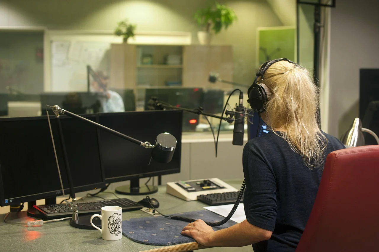 Eine Person mit langen Haaren sitzt in einem Radiostudio. Sie sitzt an einem Schreibtisch und trägt Kopfhörer.